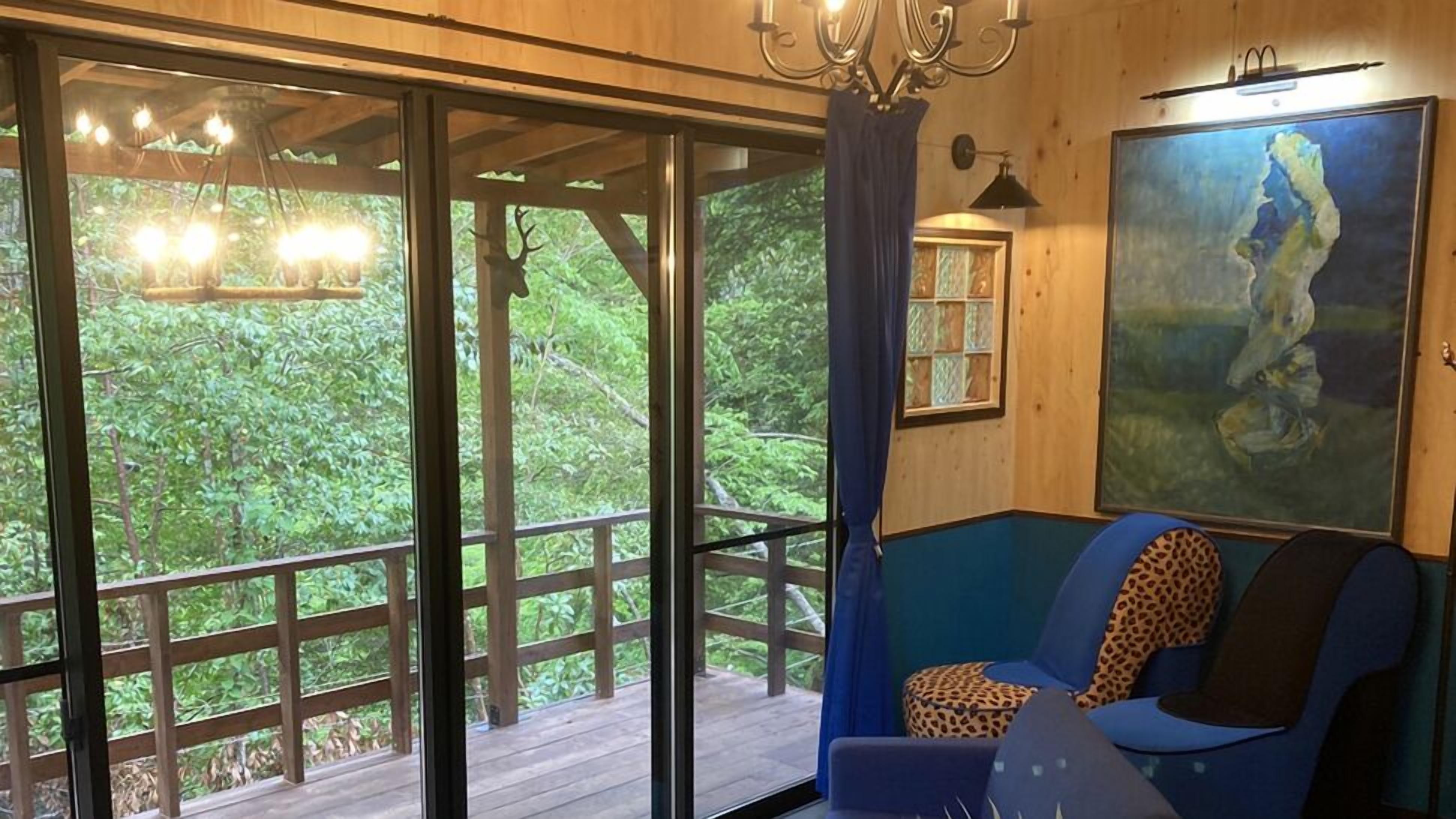 Una habitació amb una finestra gran, un sofà blau, un coixí de pell de llebrera, un quadre i una làmpada penjant.