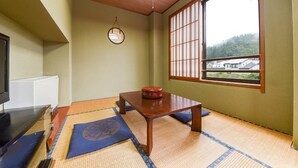 Una habitación japonesa tradicional con suelo de tatami, una mesa de madera baja, un televisor y vista a un área boscosa a través de un celosía shoji.