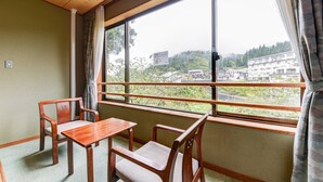 Una habitación con una mesa y sillas de madera, una ventana con vista a edificios y árboles, y una cortina.