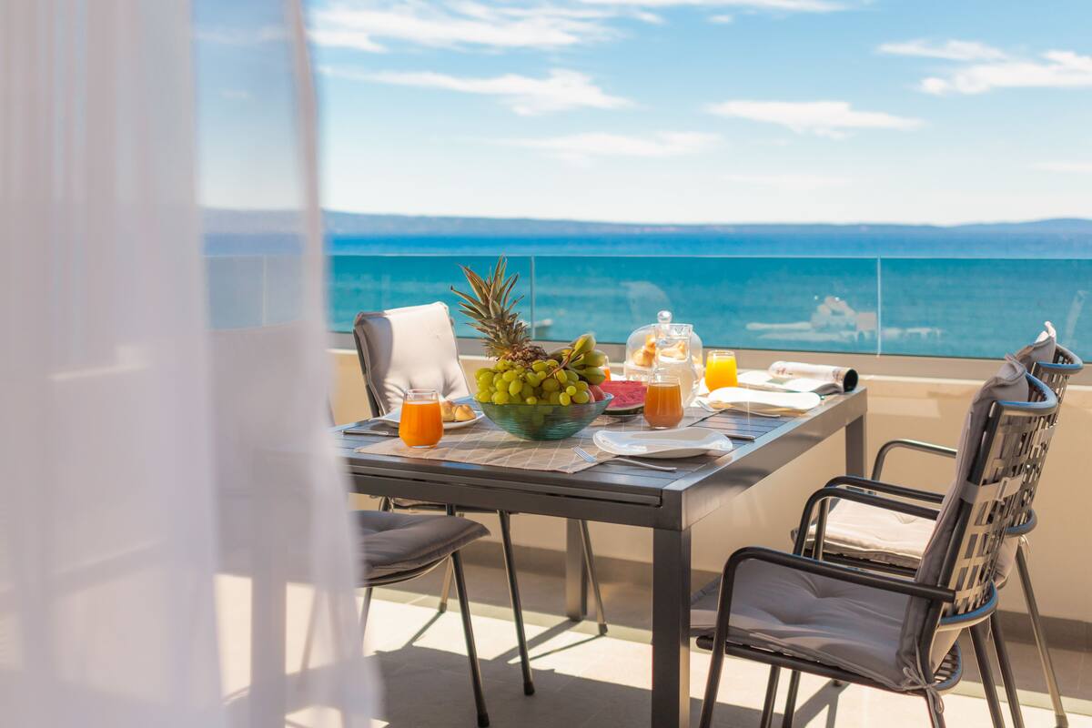 Une table sur le balcon, avec des aliments pour le petit-déjeuner, une vue sur la mer et un ciel dégagé.