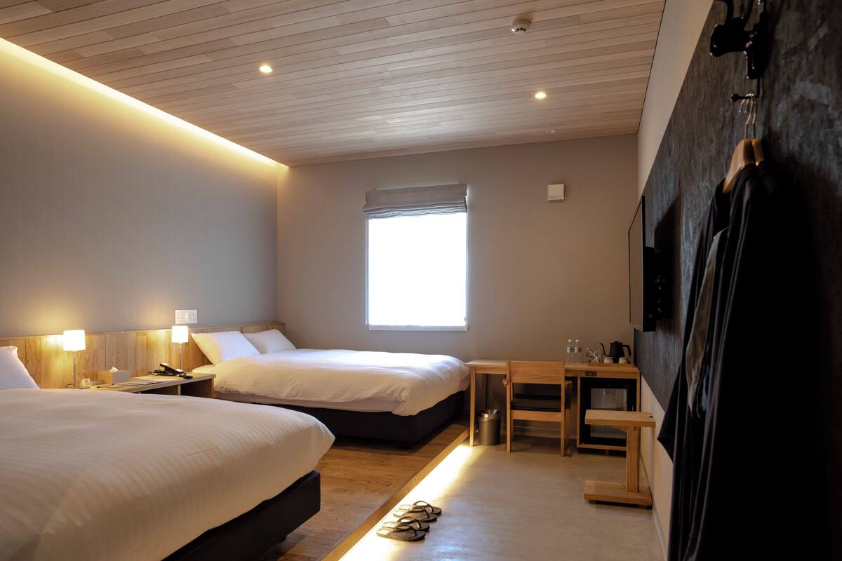 A hotel room with two beds, a small table, a chair, a window, and a wall-mounted TV.