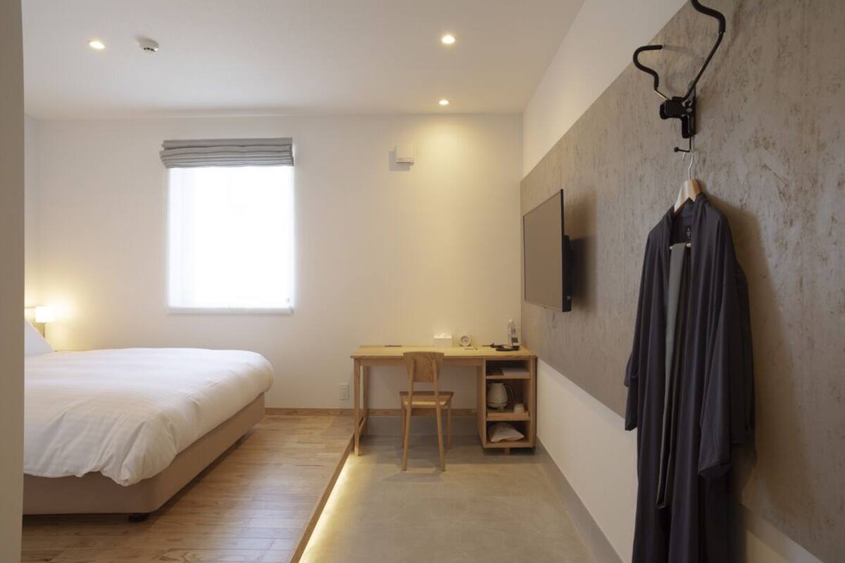 A bedroom with a bed, a desk, a chair, a TV mounted on the wall, and a coat rack with a coat.