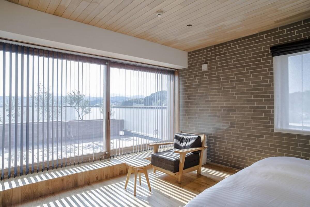 A modern bedroom with a large window, a wooden ceiling, and a brick wall.