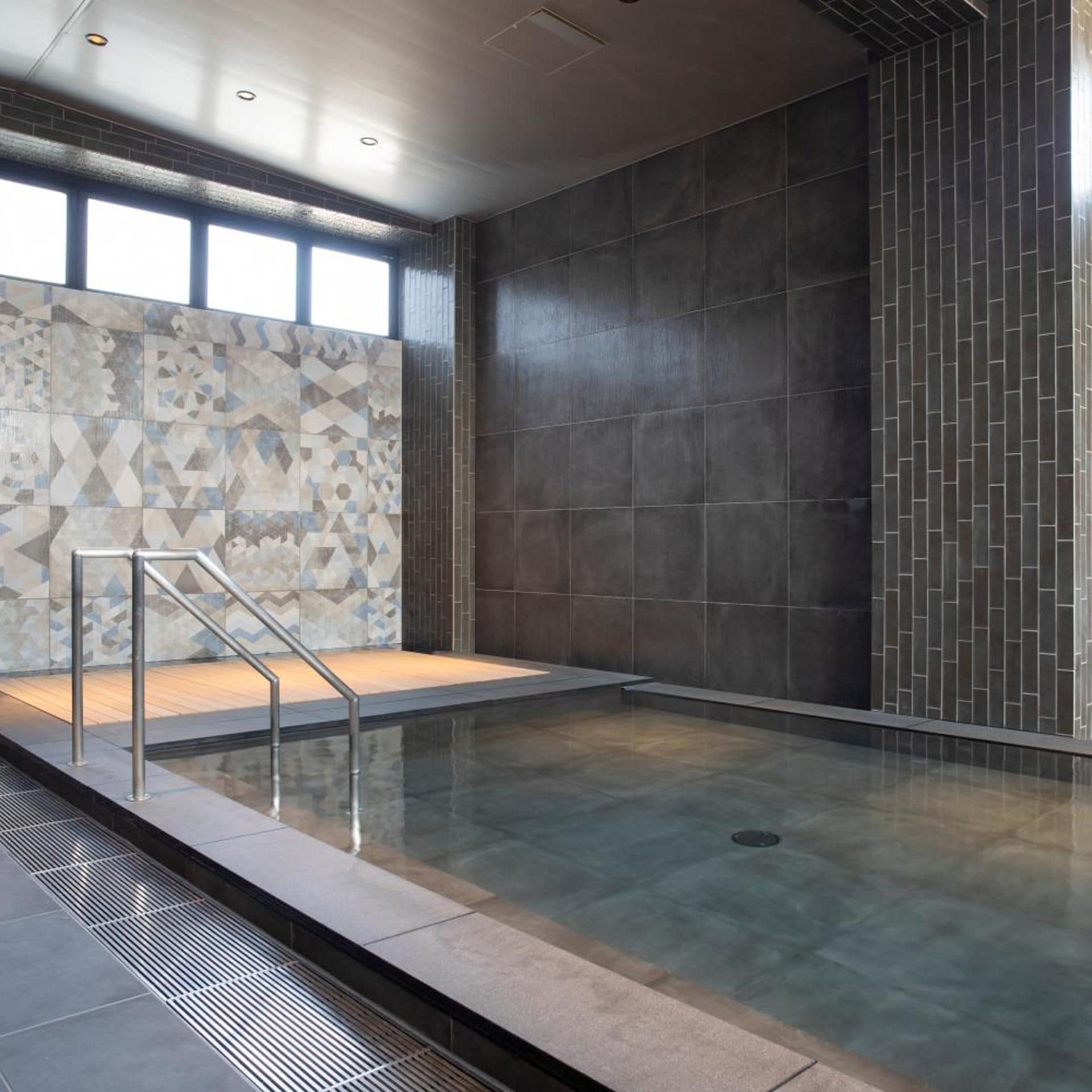 A modern indoor swimming pool with a geometric tile wall and a clear glass enclosure.