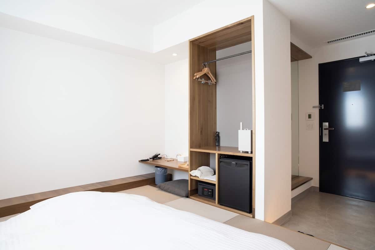 A modern hotel room with a bed, a built-in wardrobe, a small desk, and a compact kitchenette.