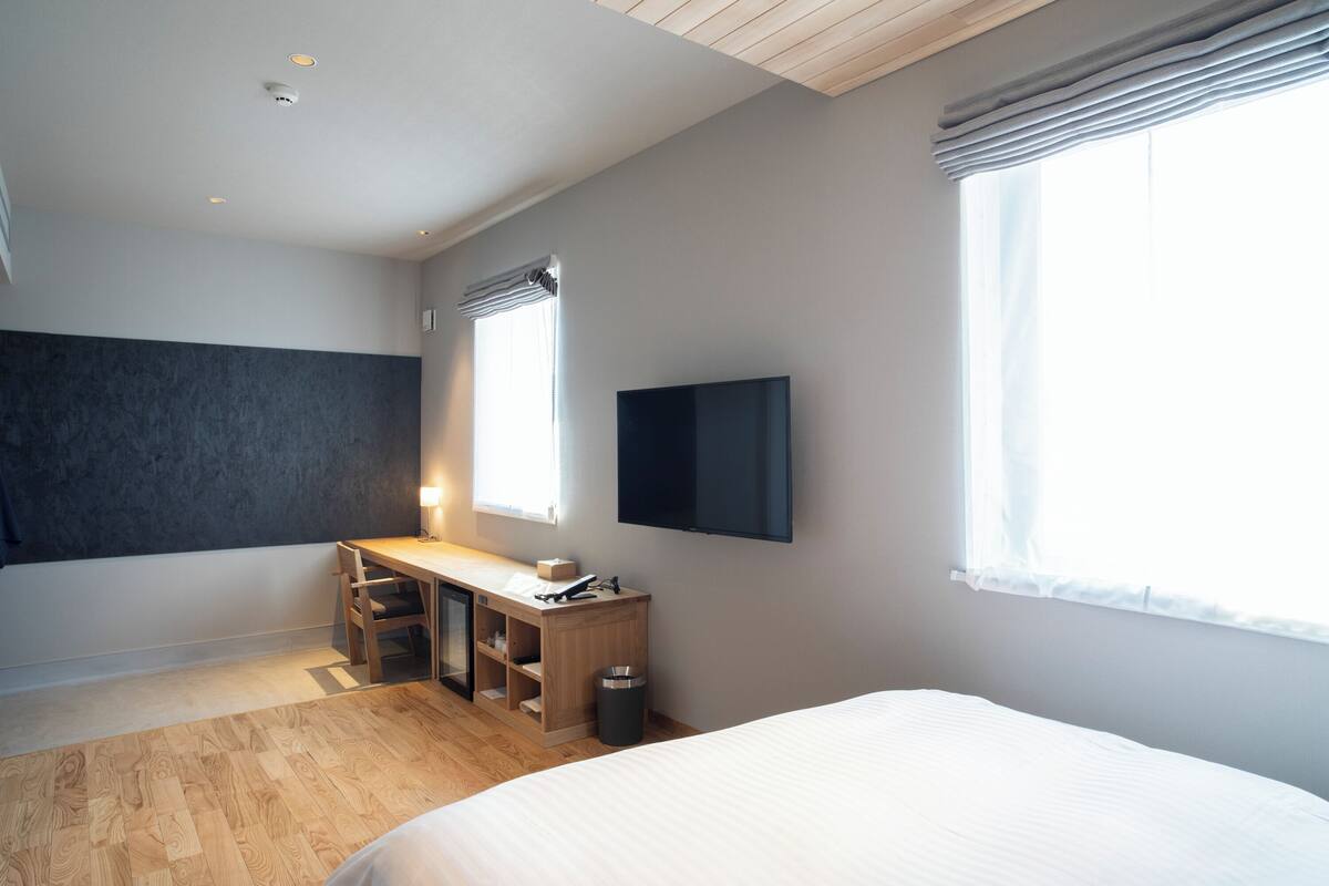 A bedroom with a bed, a desk, a chair, a TV, and a window.