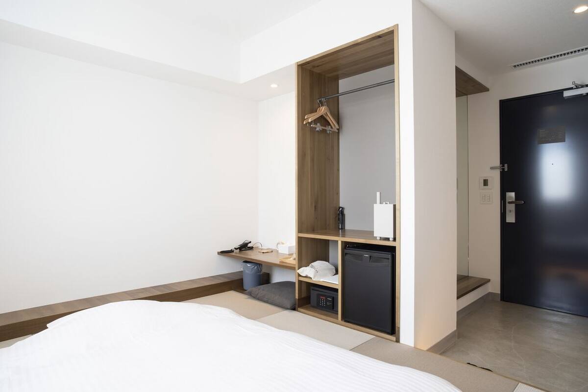 A modern hotel room with a bed, a built-in wardrobe, a small desk, and a compact kitchenette.