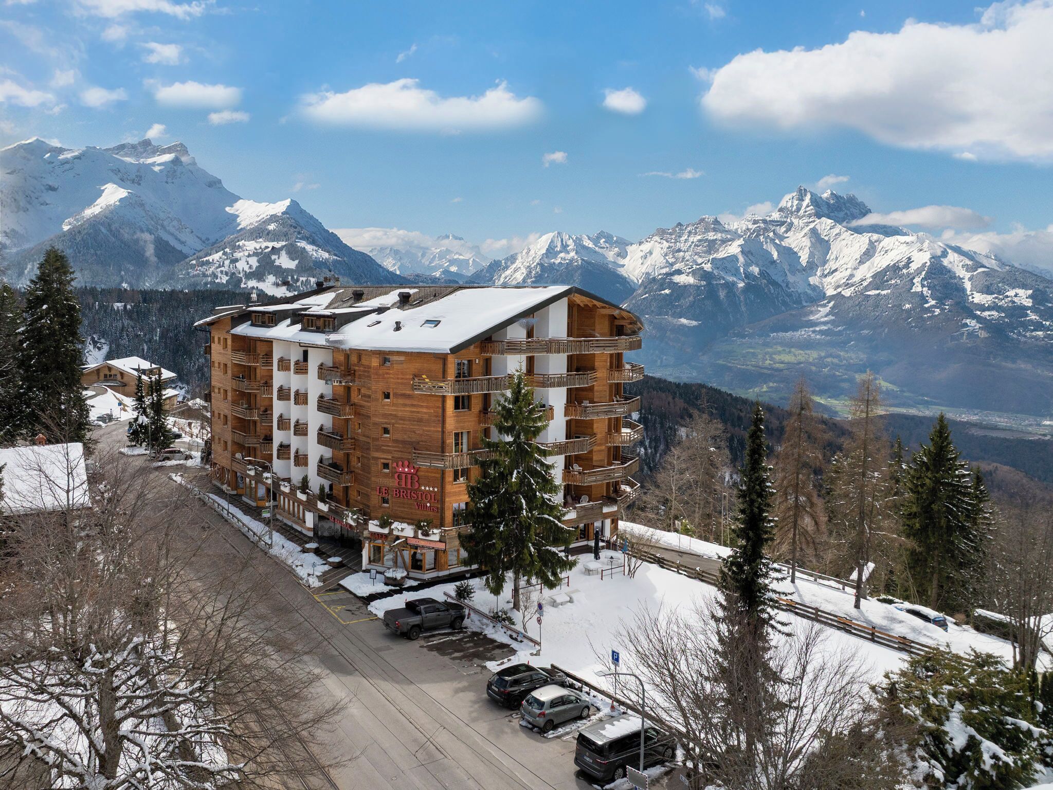 Un hotel de varios pisos con balcones, un centro de esquí al fondo y un paisaje cubierto de nieve.