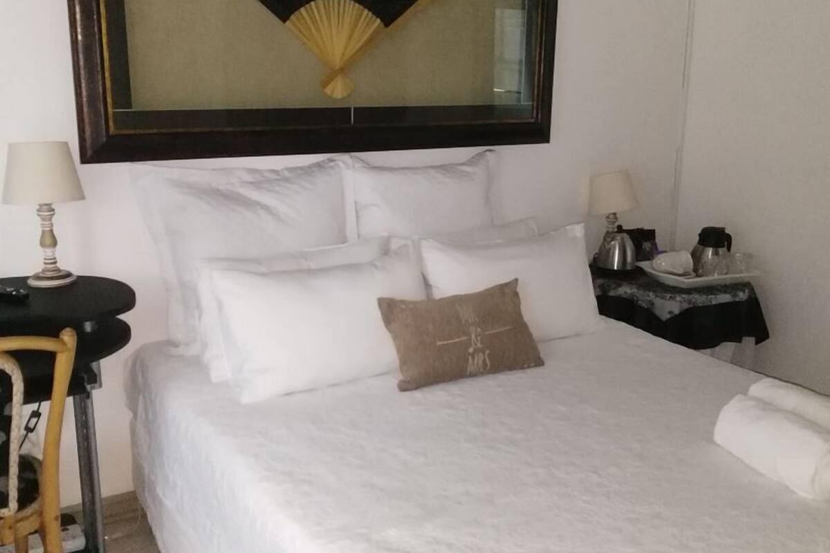 A neatly made bed with white linens and a brown pillow, a bedside table with a lamp, and a small table with a coffee pot and other items.