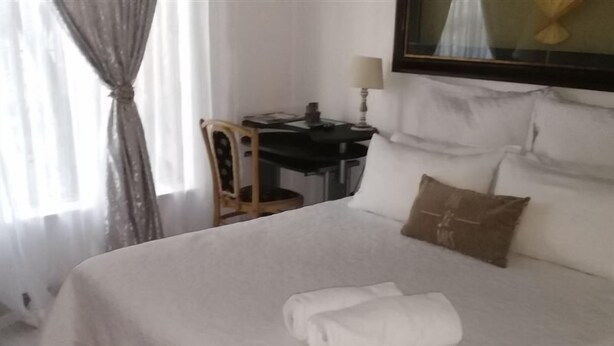 A neatly made bed with white linens and a folded towel, a small wooden chair, a desk with a lamp, and a painting on the wall. - Lapeng Home Away from Home (Maseru)
