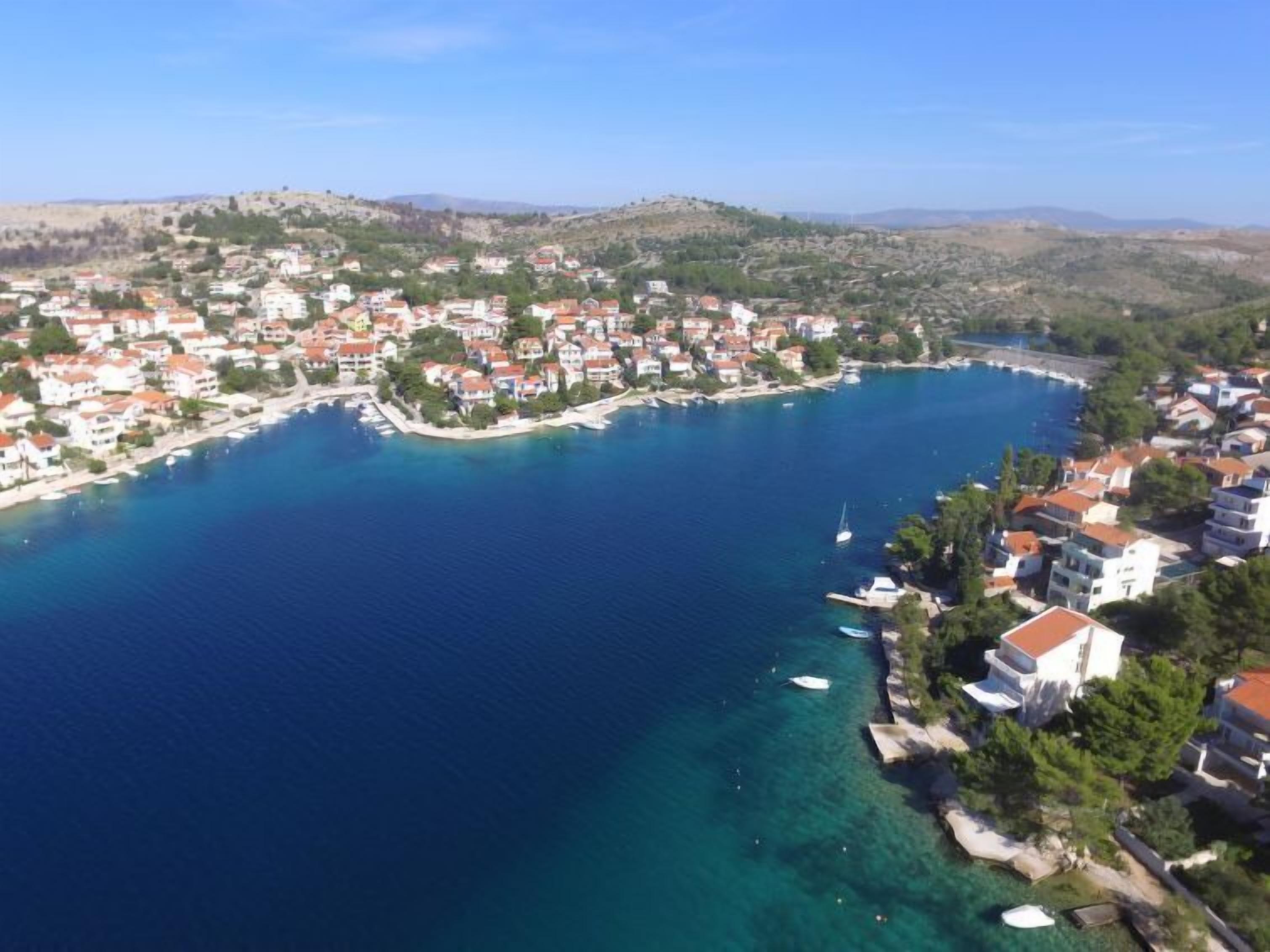 An aerial view of a coastal town with buildings along the shoreline, clear blue water, and a hilly landscape in the background.