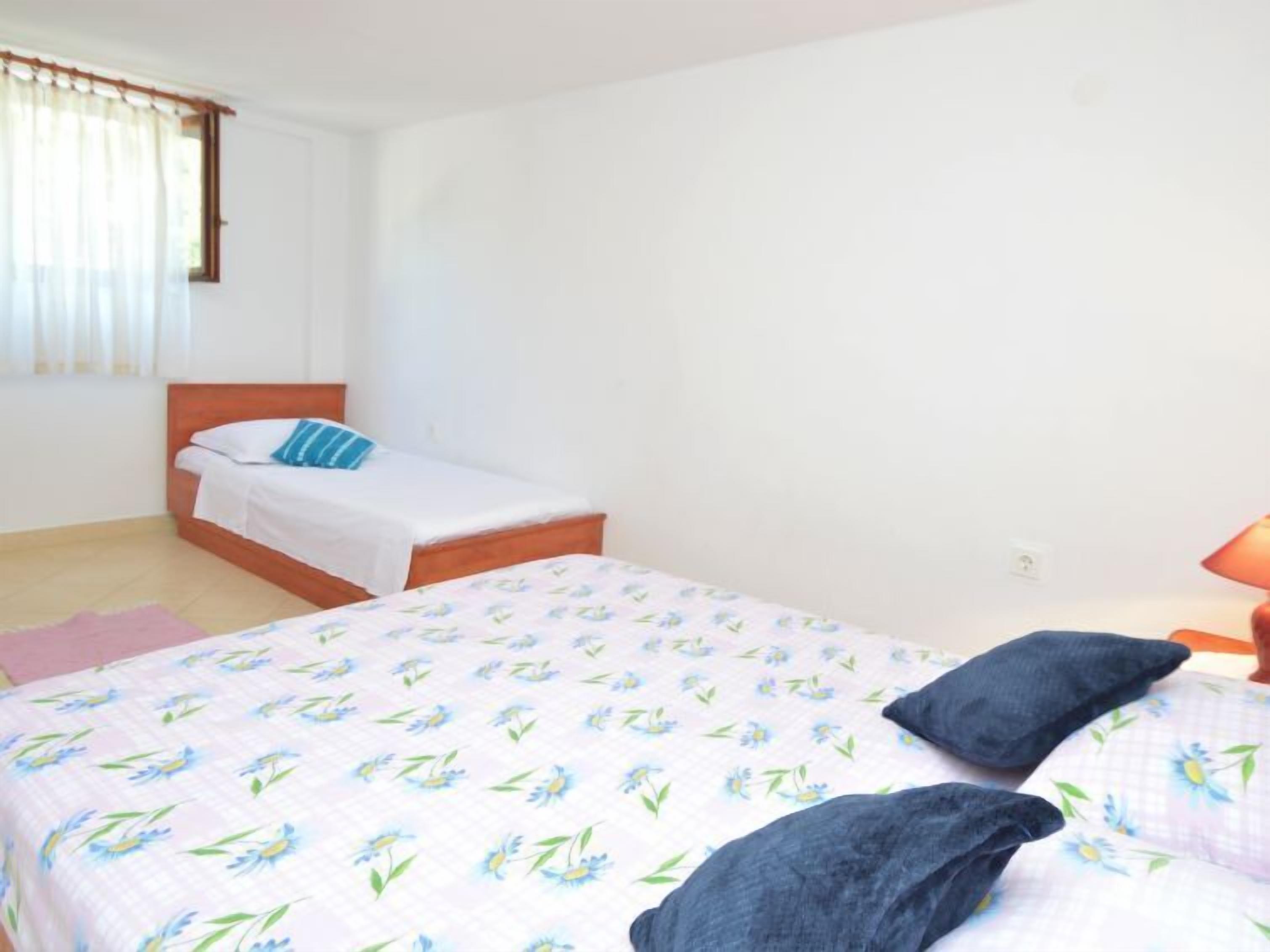 A bedroom with two beds, one with a floral bedspread and the other with a plain white bedspread. There is a nightstand with a lamp, a pink rug, and a window with curtains.