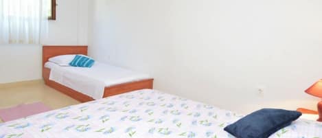 A bedroom with two beds, one with a floral bedspread and the other with a plain white bedspread. There is a nightstand with a lamp, a pink rug, and a window with curtains.