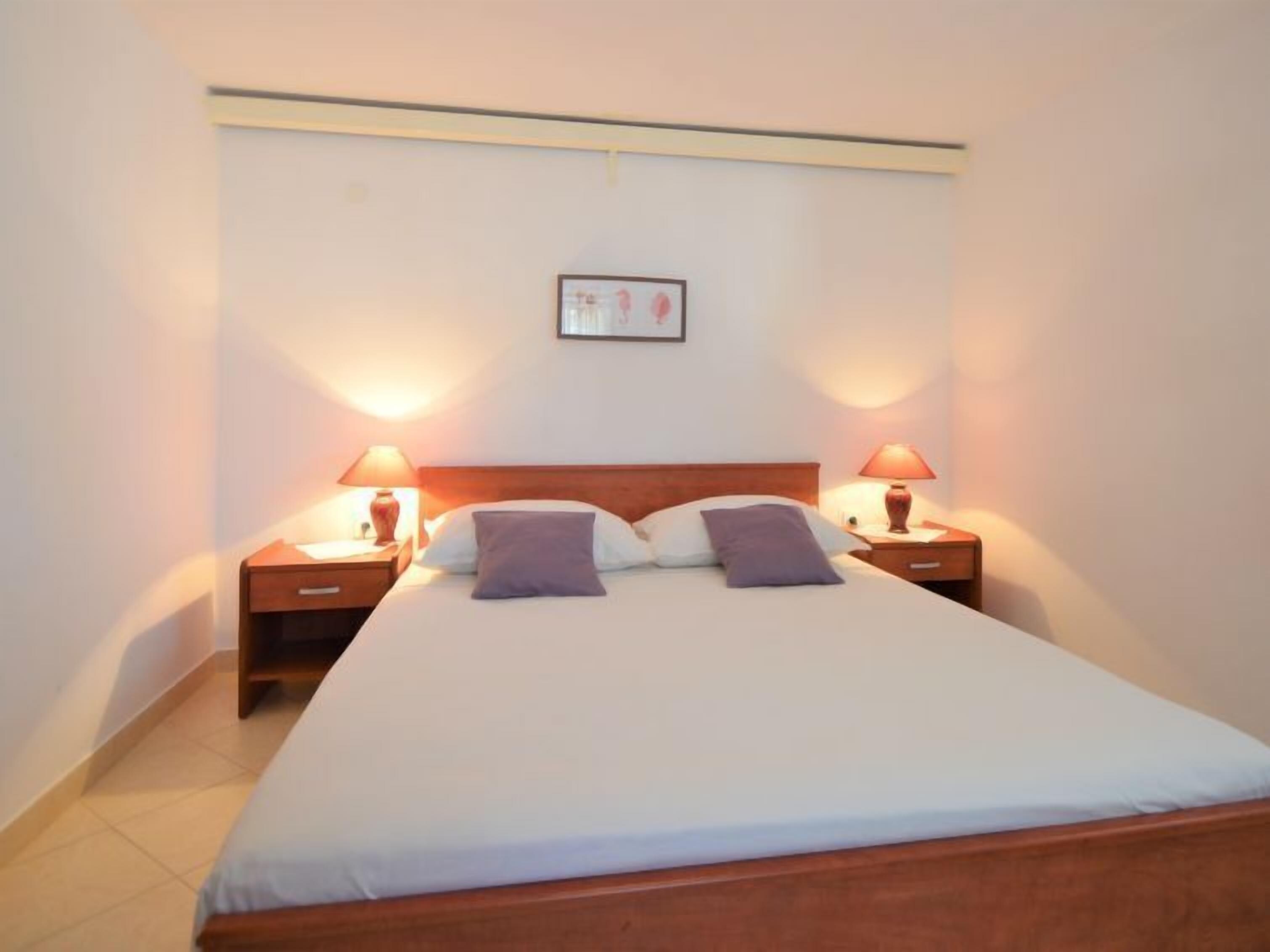 Una cama doble bien hecha, con dos almohadas, dos mesitas de noche con lámparas y un cuadro en la pared.