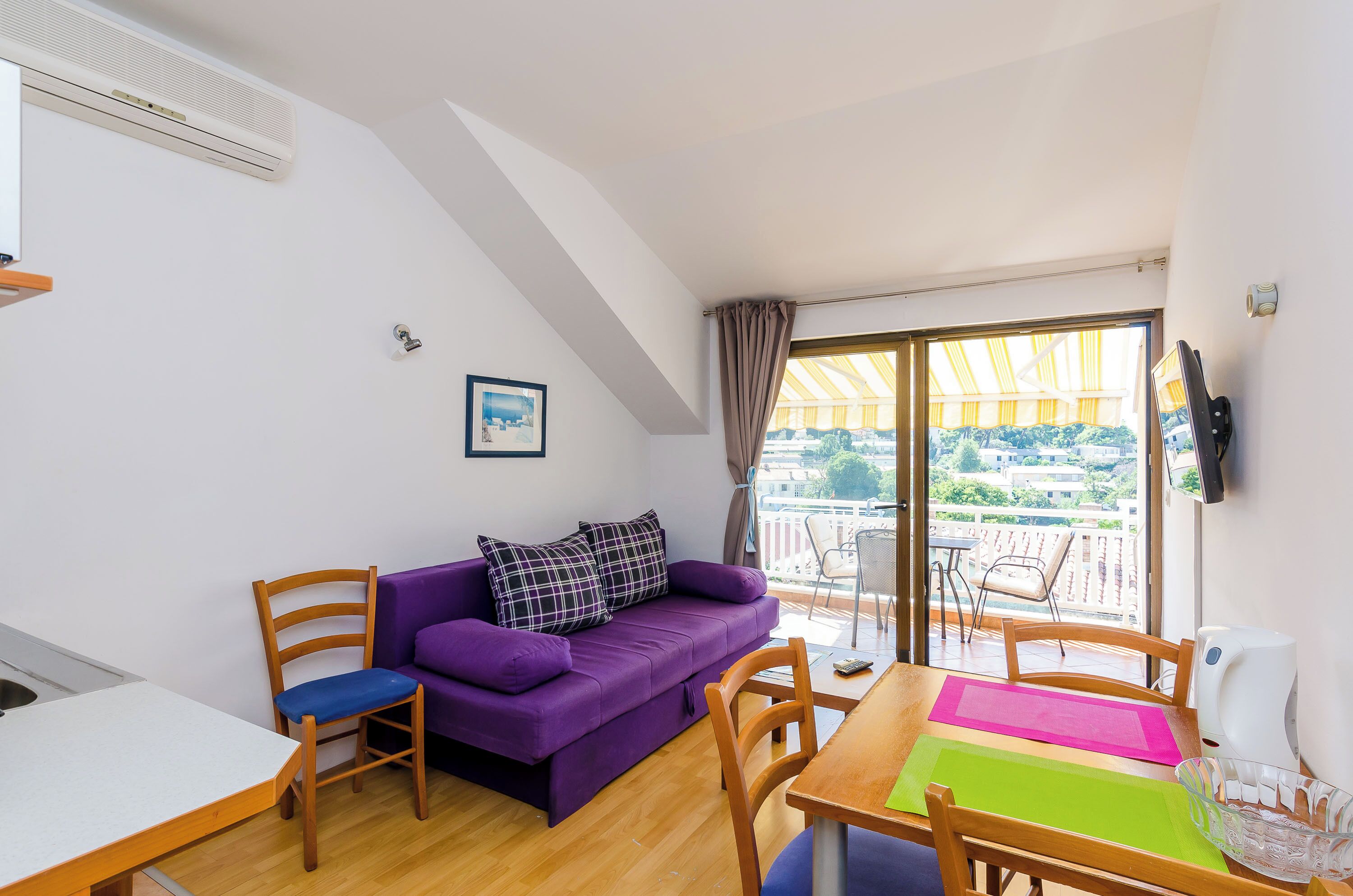 Una habitación pequeña y luminosa con un sofá morado, sillas de madera, una mesa y vistas a un balcón con barandilla y el paisaje urbano.