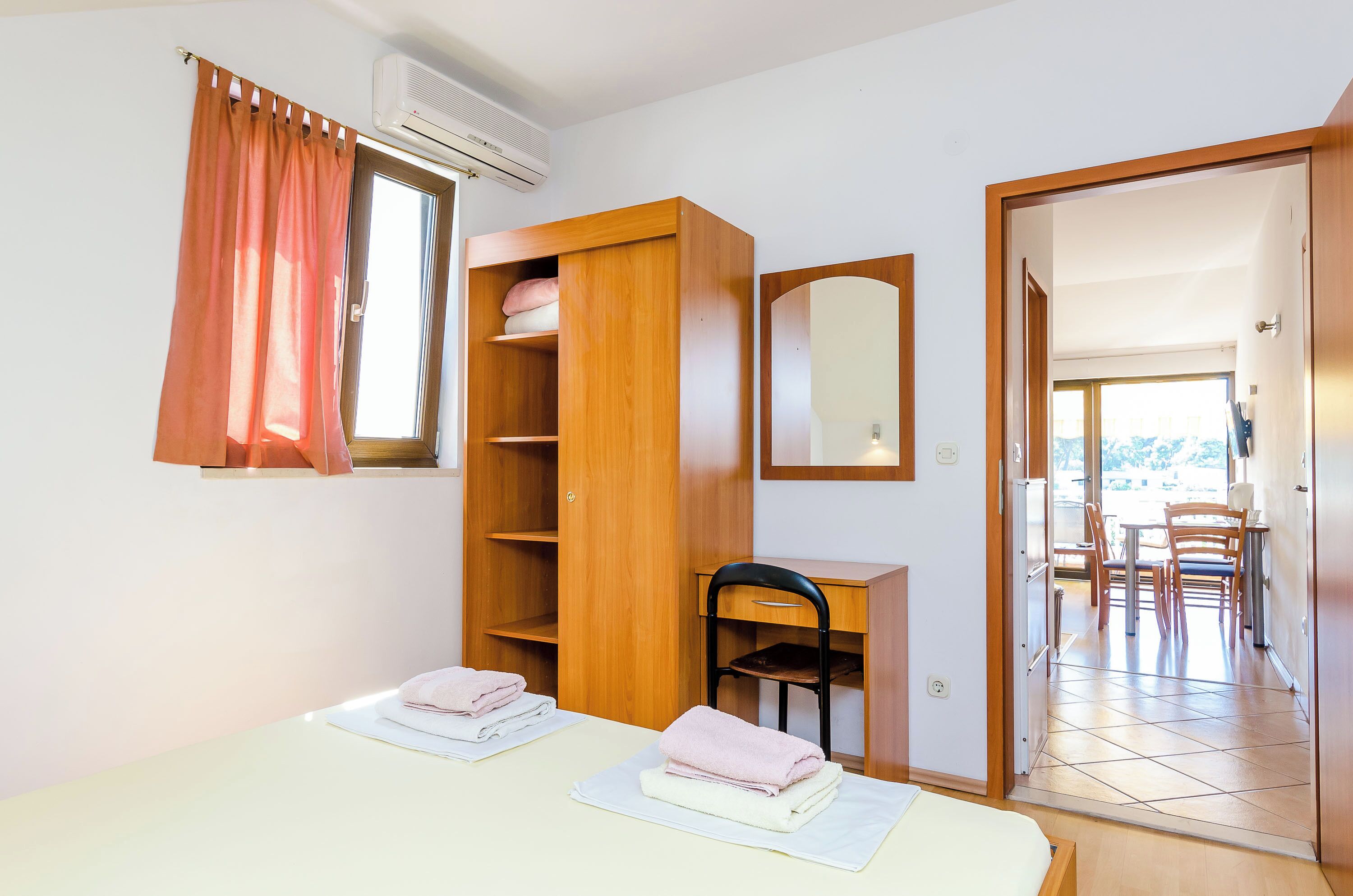 A room with a wooden wardrobe, a desk with a chair, a table with towels, and a mirror on the wall.