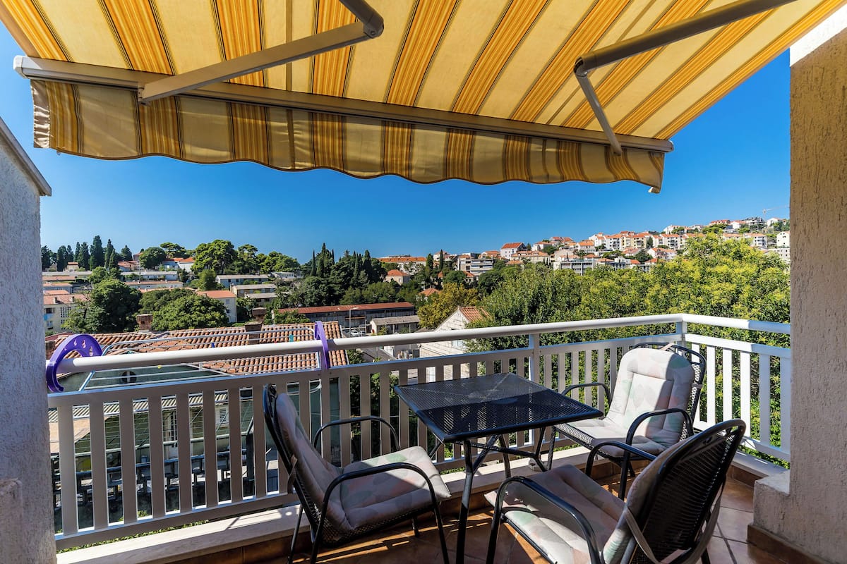Een balkon met een geel gestreept zonnescherm, tuinmeubilair en uitzicht op een stad met rode daken.