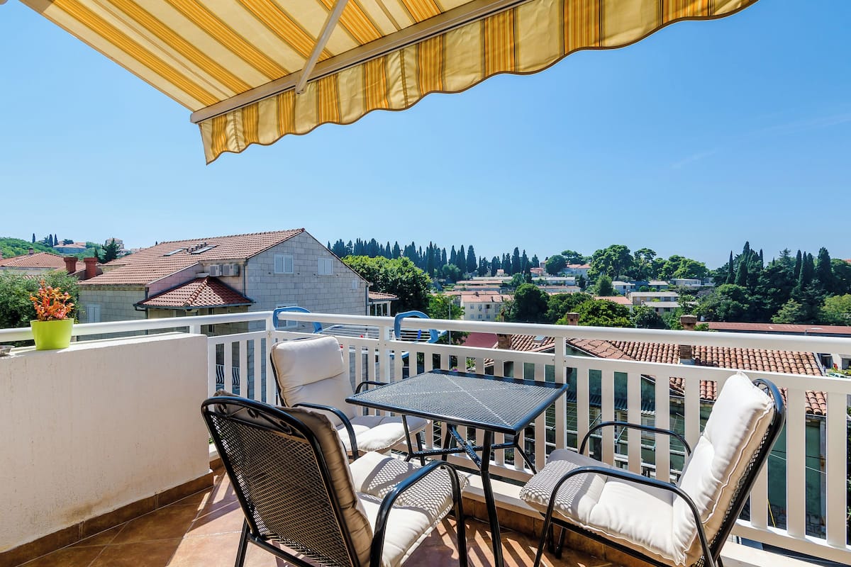 Een balkon met een tafel en stoelen, een geel zonnescherm en uitzicht op een stad met huizen en bomen.