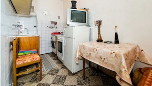 A compact kitchen with white appliances, a wooden chair, a table with a floral tablecloth, and a vase with flowers. - Rooms Kisic (Dubrovnik)
