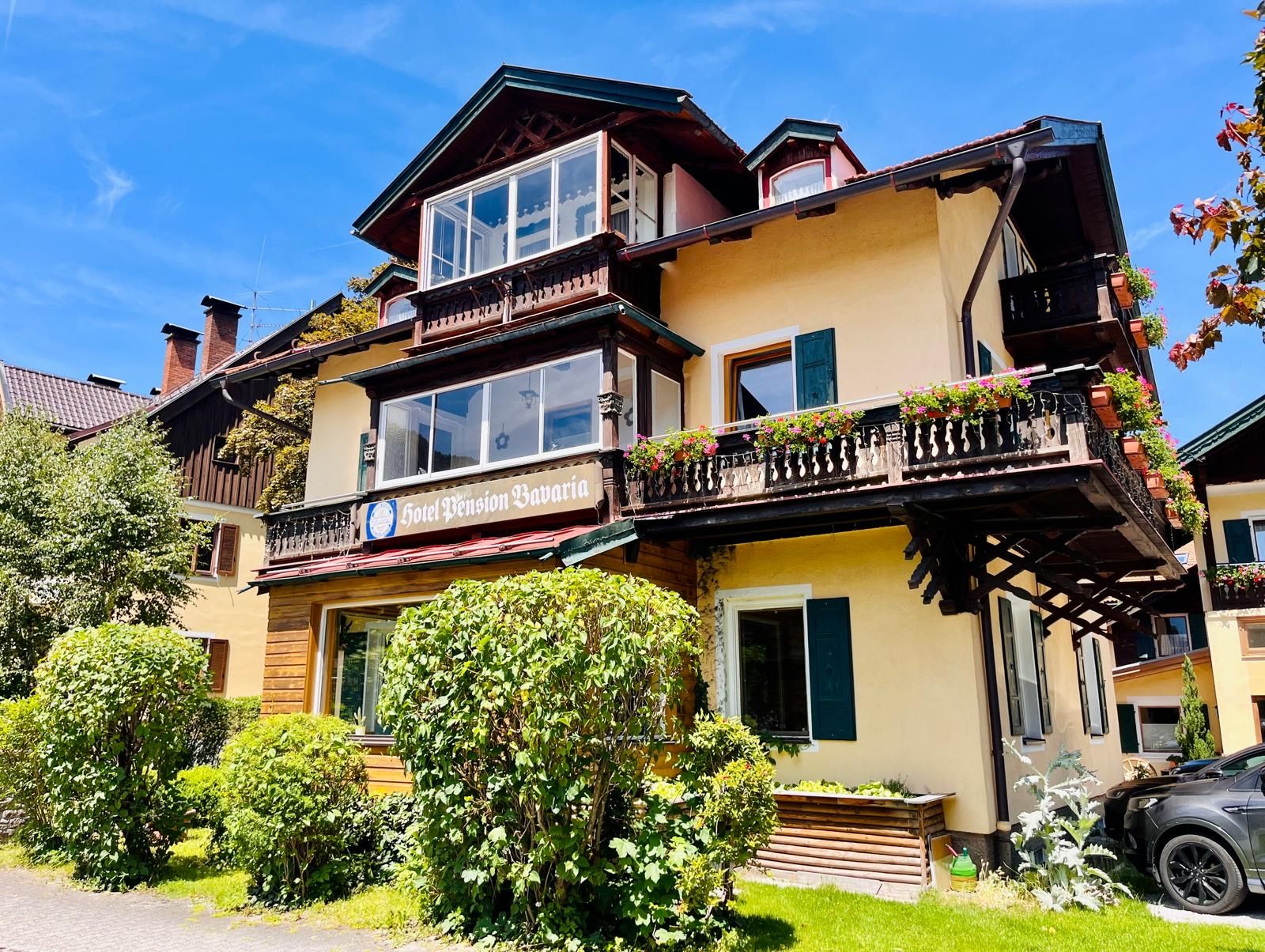 Un hôtel de deux étages avec un balcon, une enseigne indiquant « Hôtel Pension Bavaria », et une voiture garée devant.