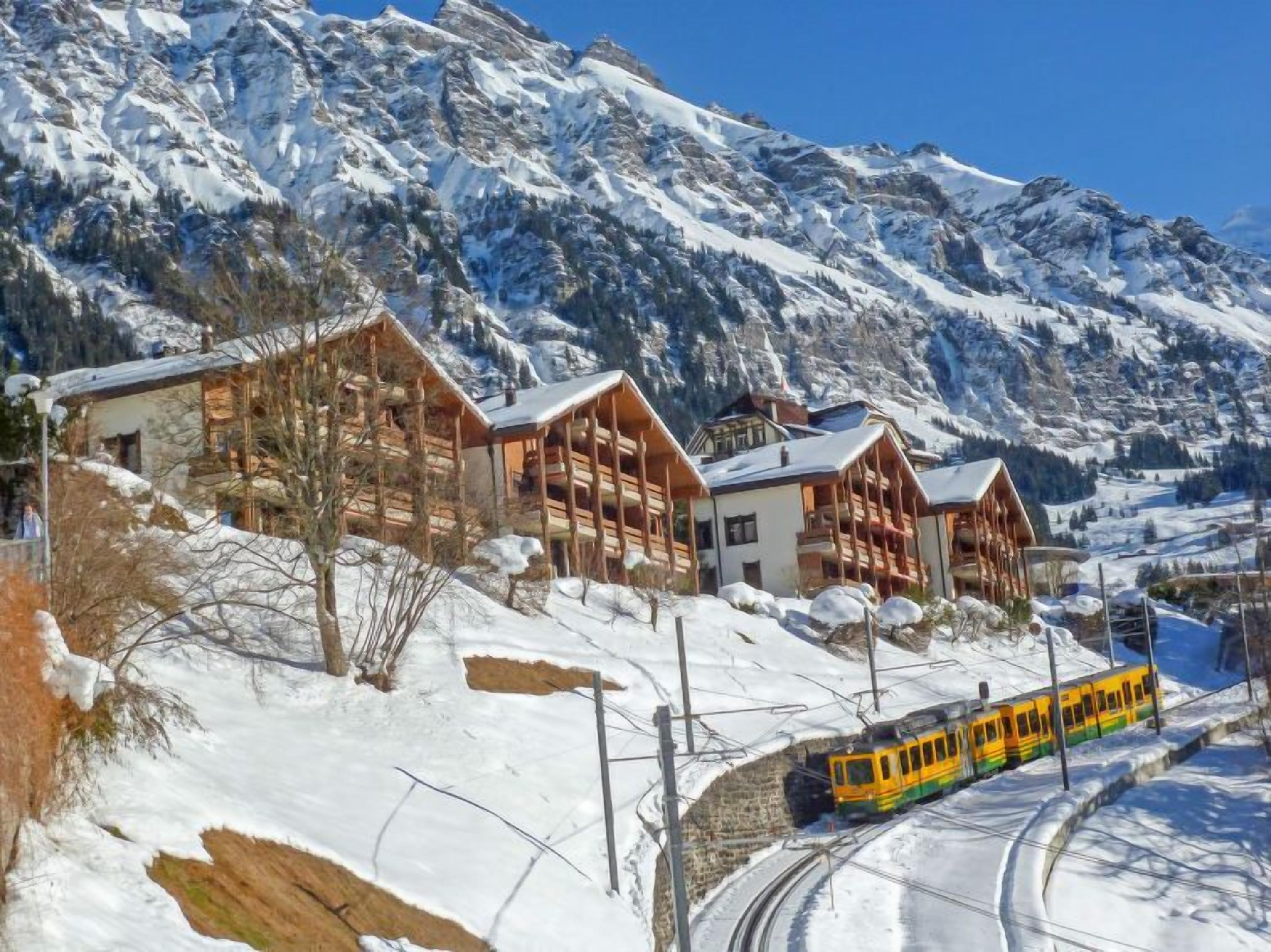 Une montagne enneigée, parsemée de chalets en bois, et un train jaune circulant sur des rails.