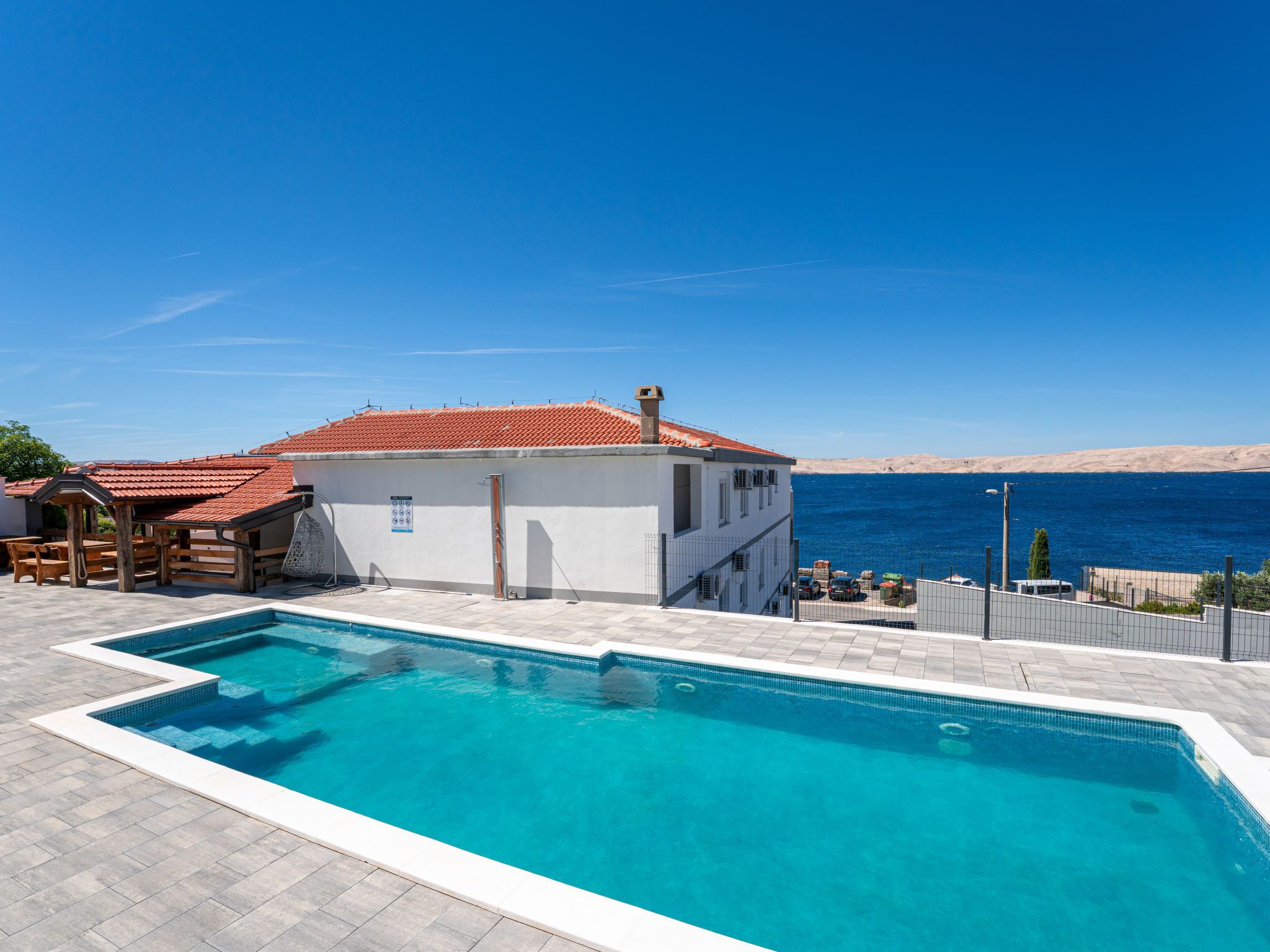 A pool area with a clear view of the sea, a building with a red-tiled roof, and a fenced patio.