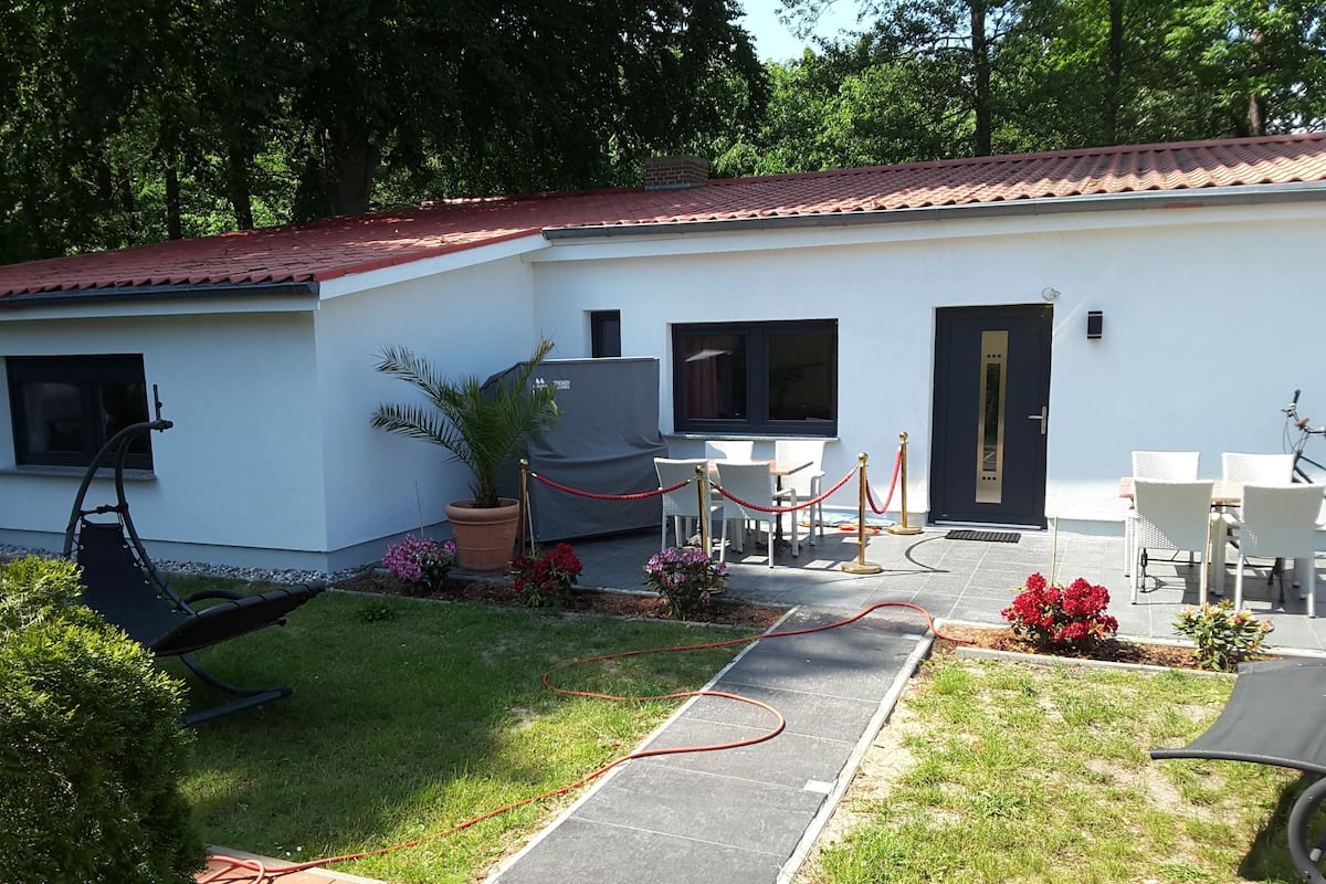 Un edificio blanco con techo de tejas rojas, un patio con muebles blancos y un jardín con plantas en macetas y una palmera.