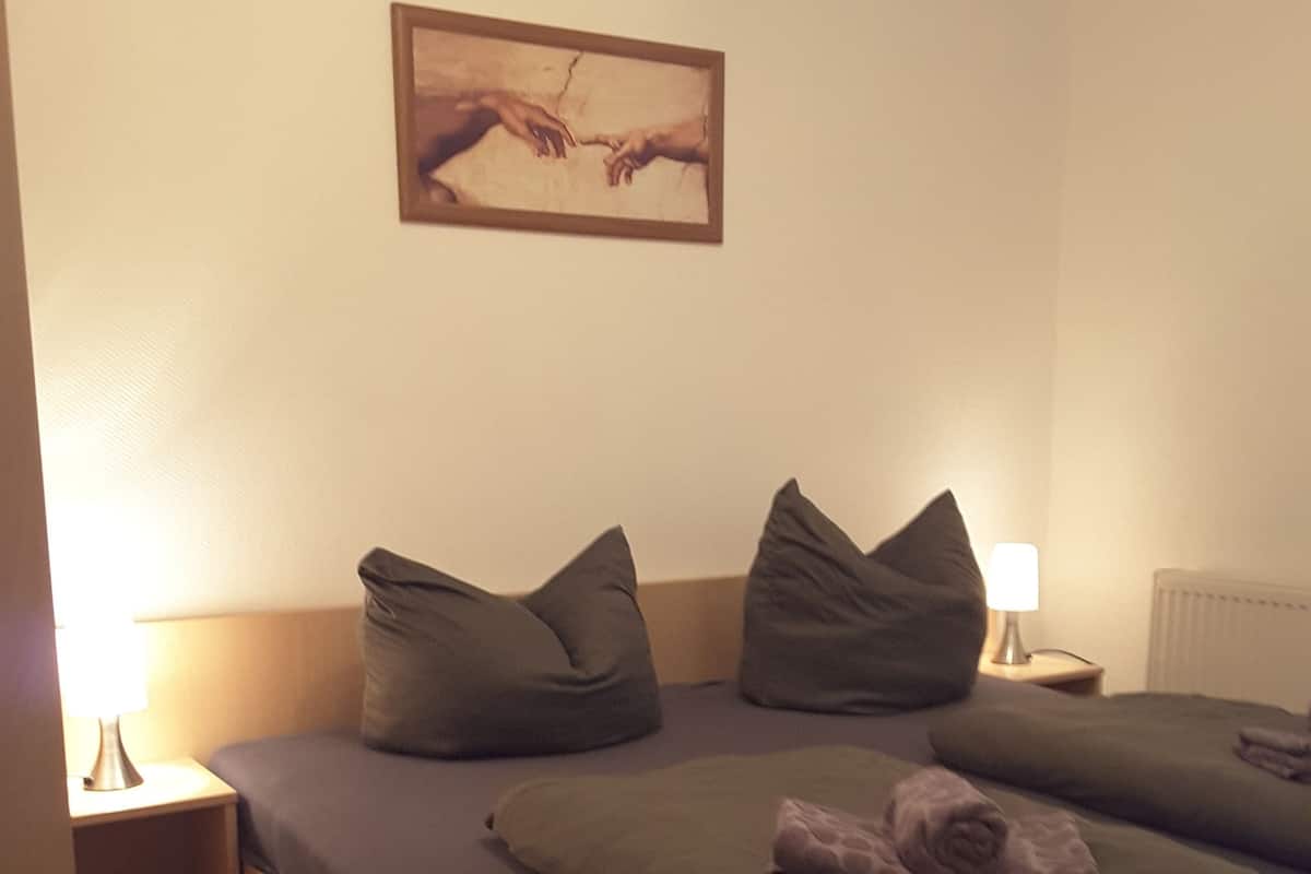 Un dormitorio con cama, almohadas, mesita de noche, lámpara y un cuadro en la pared.