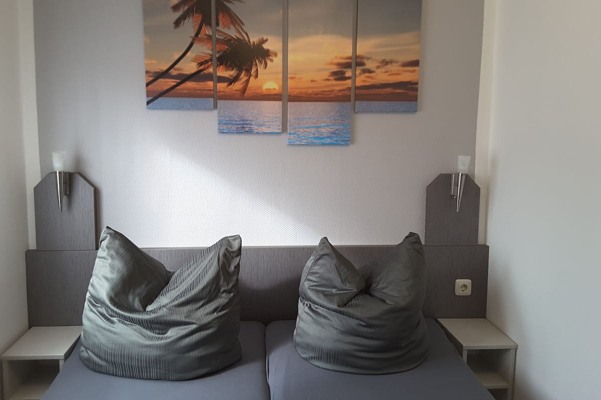 Una habitación de hotel moderna con un mural grande de un atardecer sobre el océano, dos mesitas de noche con lámparas y dos sillones tipo bean bag grises.