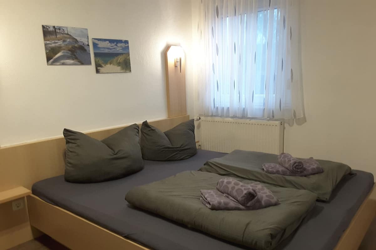 Una cama individual con estructura de madera, ropa de cama gris y una ventana con cortinas traslúcidas.