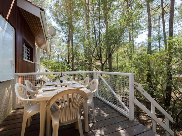 A wooden deck with a table set for dining, surrounded by white plastic chairs, overlooking a forested area.
