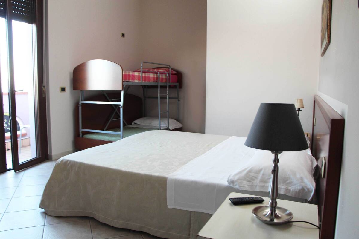 Ein Hotelzimmer mit einem Einzelbett, einem Schreibtisch mit Lampe, einem Sessel und einem Stockbett, sowie einer Tür zu einem Balkon.