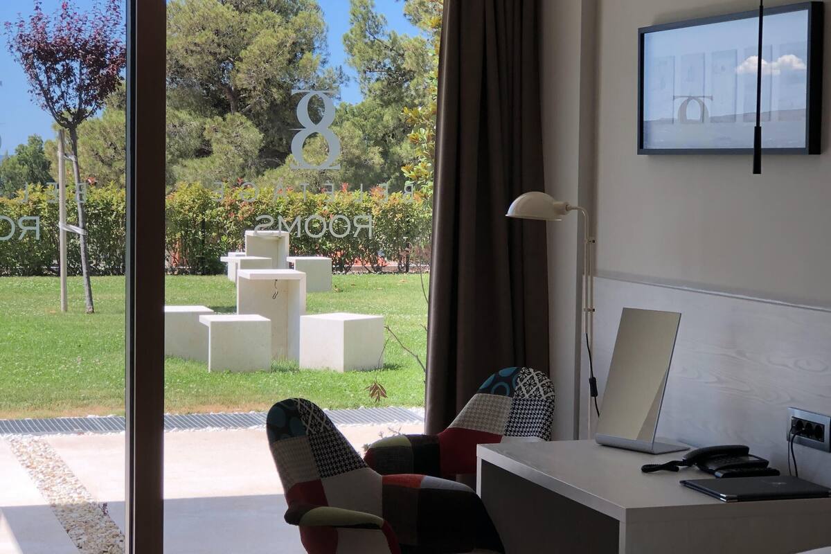 A modern hotel room with a bed, a desk with a laptop, and a view of a garden through a glass door.