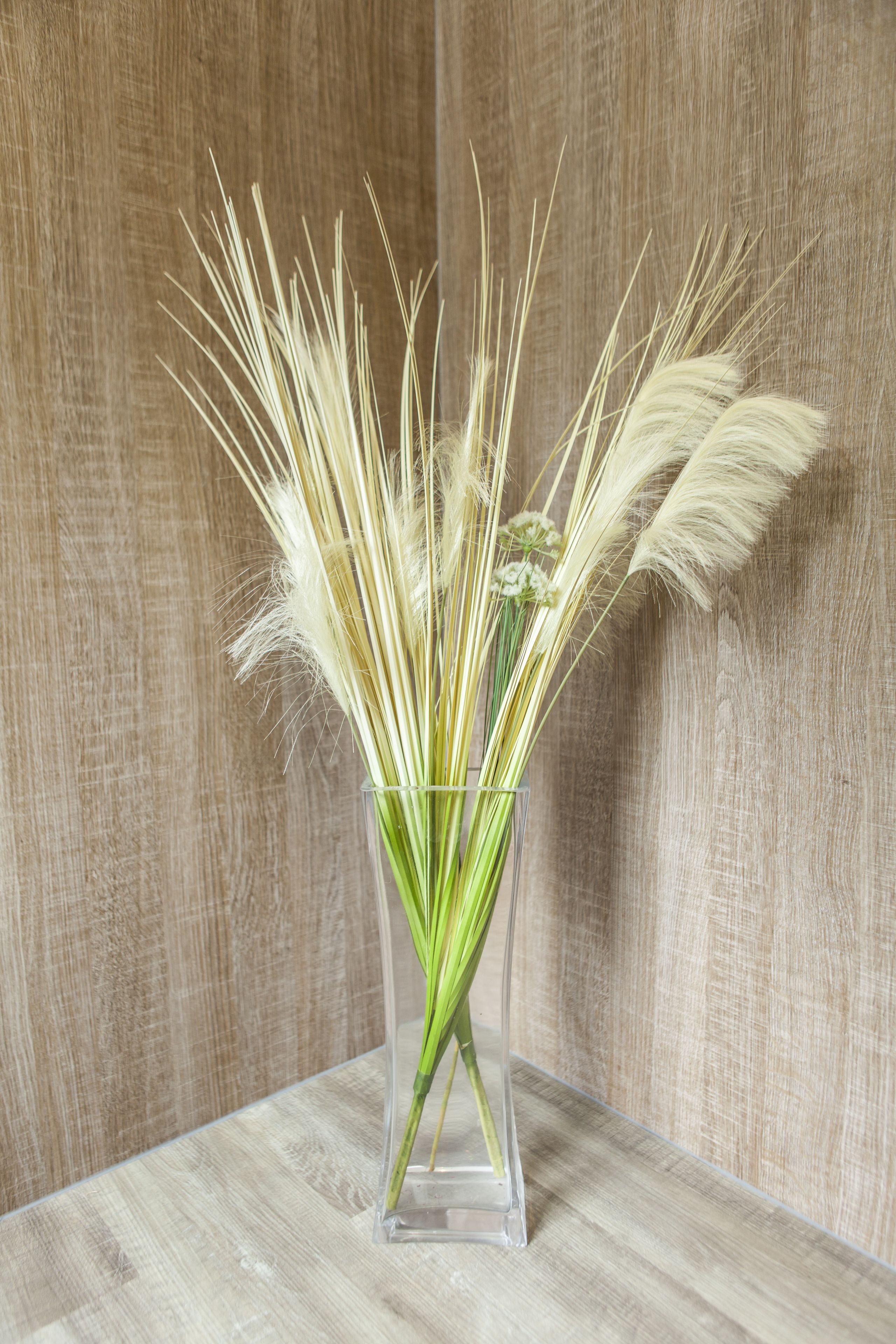 A clear glass vase with long, slender green stems and fluffy white grass-like elements, placed on a wooden surface against a textured wall.