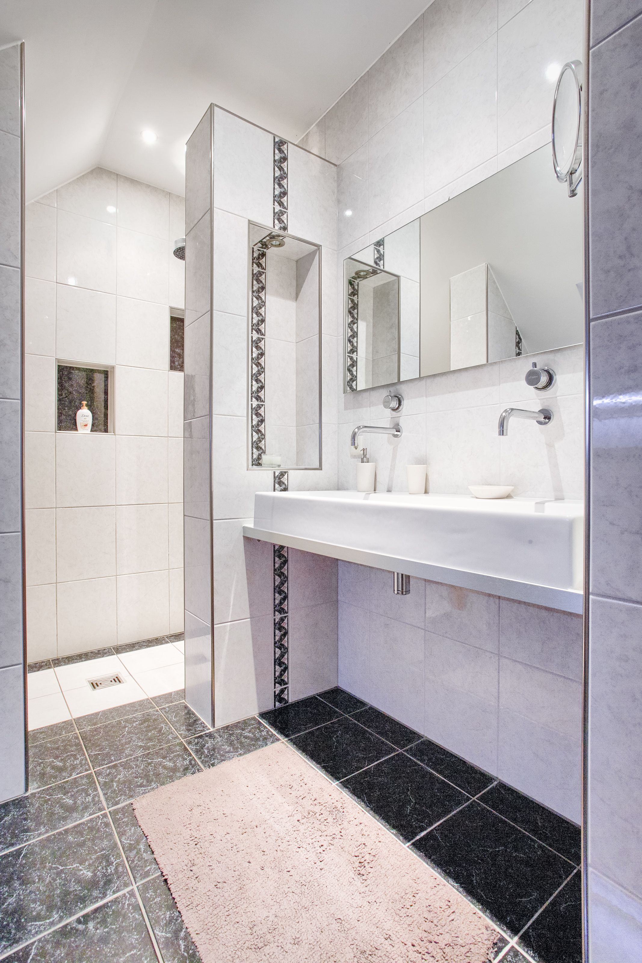Un baño moderno con ducha envuelta en vidrio, tocador blanco con un espejo grande y suelo de baldosas blanco y negro.