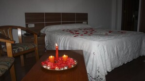 Habitación de hotel con cama, mesita de noche con velas y pétalos de rosa, y una silla de madera.