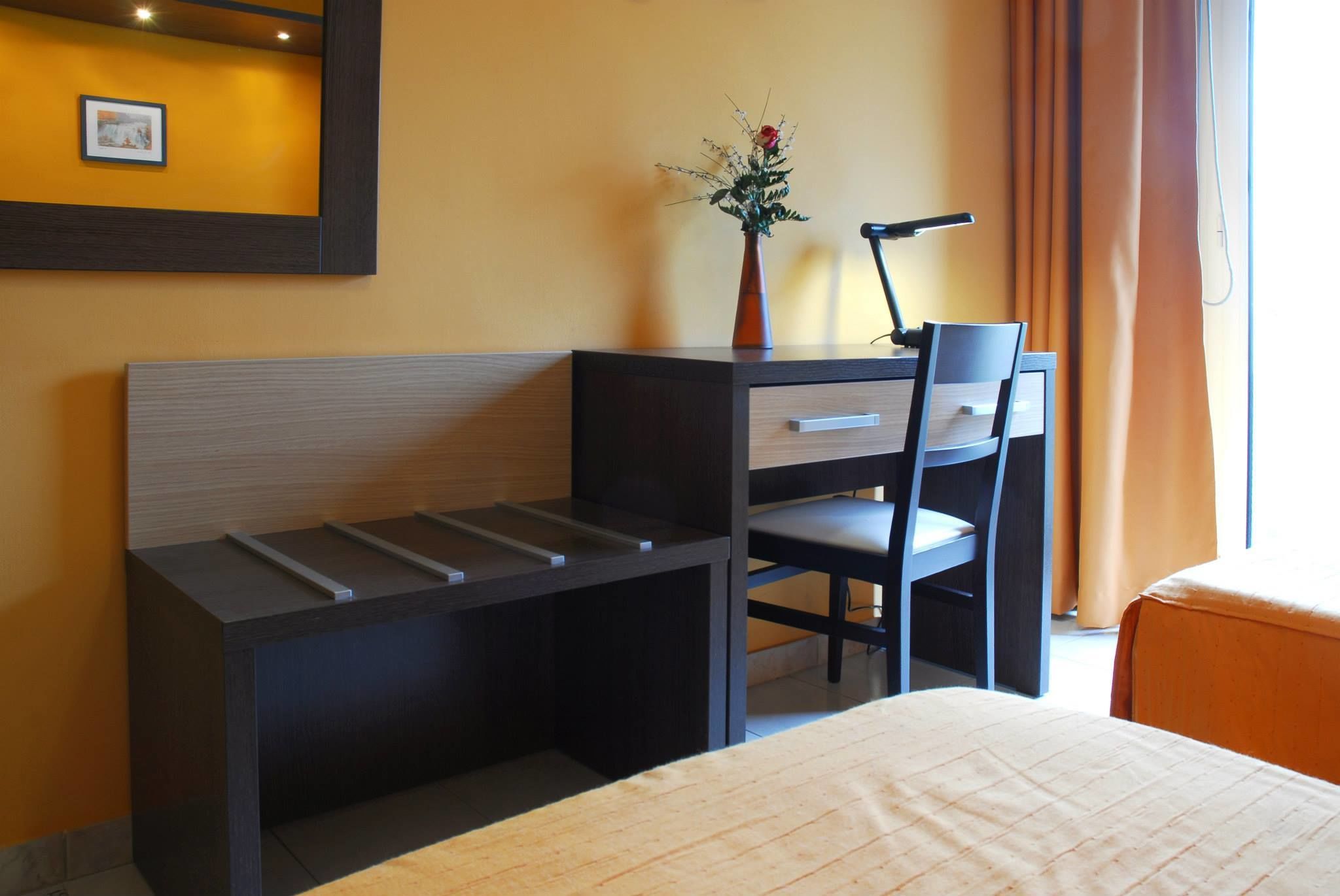 Habitación de hotel con cama, escritorio con silla, una mesita, un florero con flores y un cuadro en la pared.