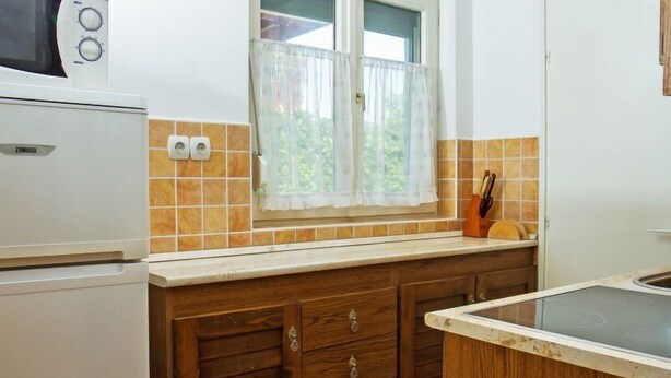 A kitchen with a microwave, a window with curtains, a tiled backsplash, wooden cabinets, and a countertop. - Apartment Vilma by Interhome (Balatonmariafurdo)