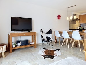 A modern living room with a flat-screen TV, a wooden entertainment unit, a cowhide rug, and a dining area with white chairs and a wooden table.
