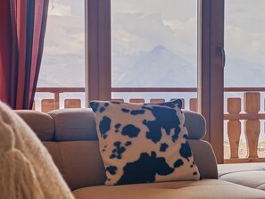 A sofa with a cow-patterned pillow, a wooden railing, and a mountain view through large windows.