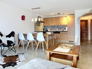 A modern living room with a dining area, a kitchen with wooden cabinets, and a hallway leading to a door.
