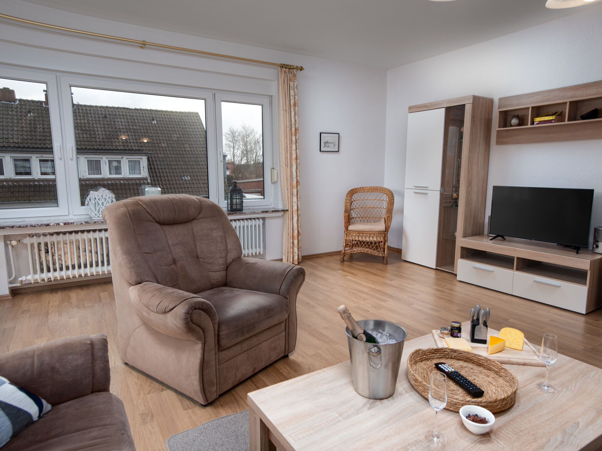 Ein modernes Wohnzimmer mit Ledersofa, Korbstuhl, Fernseher auf Holzständer und Esstisch mit Champagner und Käse.