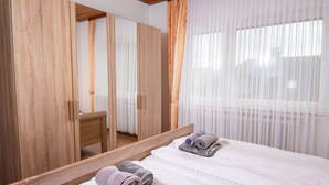 Ein Hotelzimmer mit Holzschrank, Bett mit weißer Bettwäsche und großem Fenster mit durchsichtigen Vorhängen.