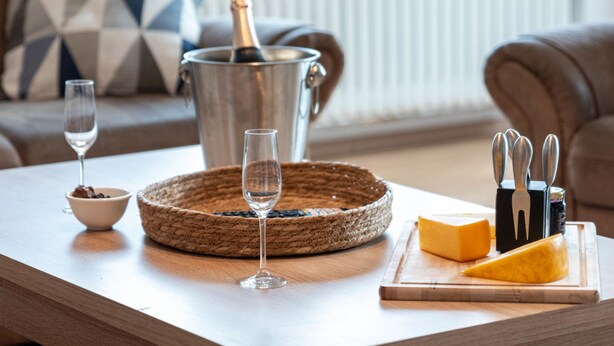 Ein Holztisch mit einer Champagnerflasche, zwei Weingläsern, einer Schale Schokolade und einer Käseplatte mit Besteck.
