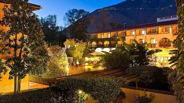 Ein Hotelinnenhof im Abenddämmerung, mit Blick auf einen Berg, mit einem gut beleuchteten Außenbereich, Pflanzen und einer überdachten Sitzgelegenheit.