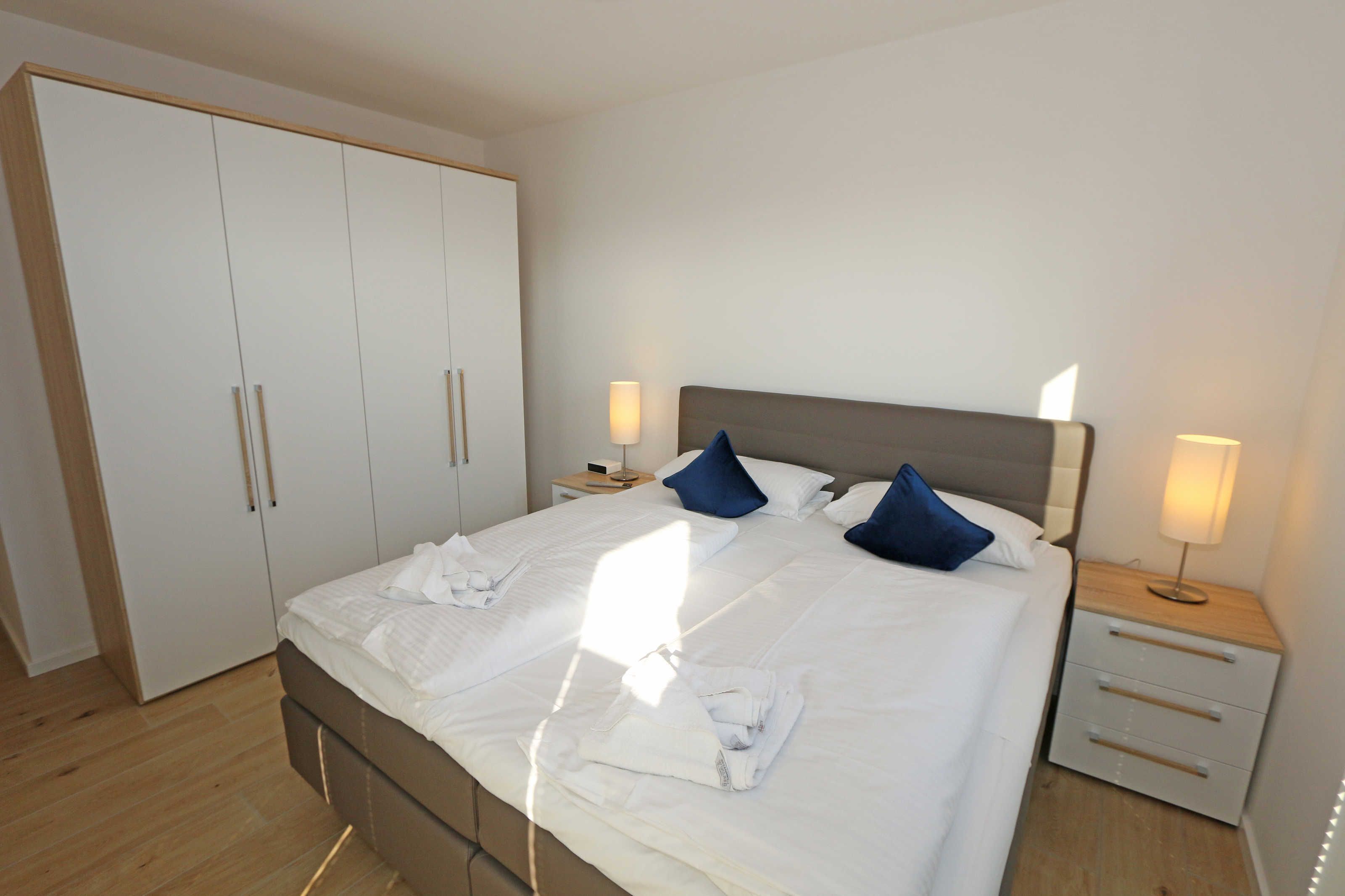 Ein ordentlich bezogenes Bett mit Kissen, ein Nachttisch mit Lampe und ein Kleiderschrank.