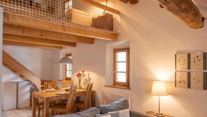 Ein gemütliches Zimmer mit Holzbalkendecke, einem Esstisch mit Stühlen, einem grauen Sofa und einer Lampe.