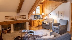 Ein gemütlicher Loftraum mit freiliegenden Holzbalken, einem Kamin, einem Schreibtisch mit Computer, einem bequemen Sessel und einer Couch.