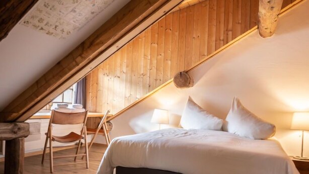 Ein gemütliches Schlafzimmer im Dachgeschoss mit holzgetäfelter Schräge, einem Bett mit weißer Bettwäsche, einem Sessel und einem kleinen Tisch.