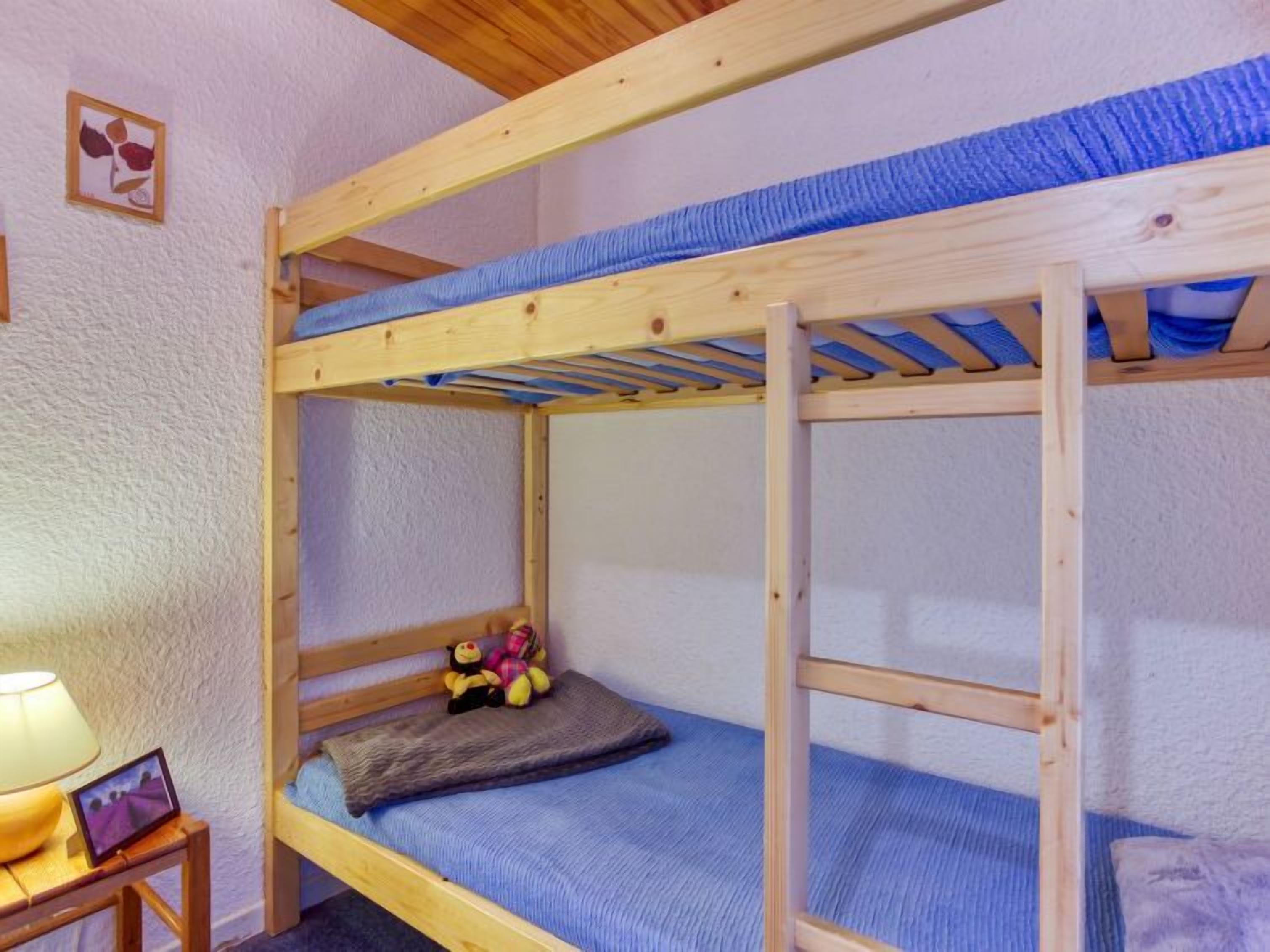 Ein Stockbett mit Holzrahmen, einer blauen Matratze und einer Leiter. Links daneben steht ein kleiner Tisch mit einer Lampe und einem Bilderrahmen.